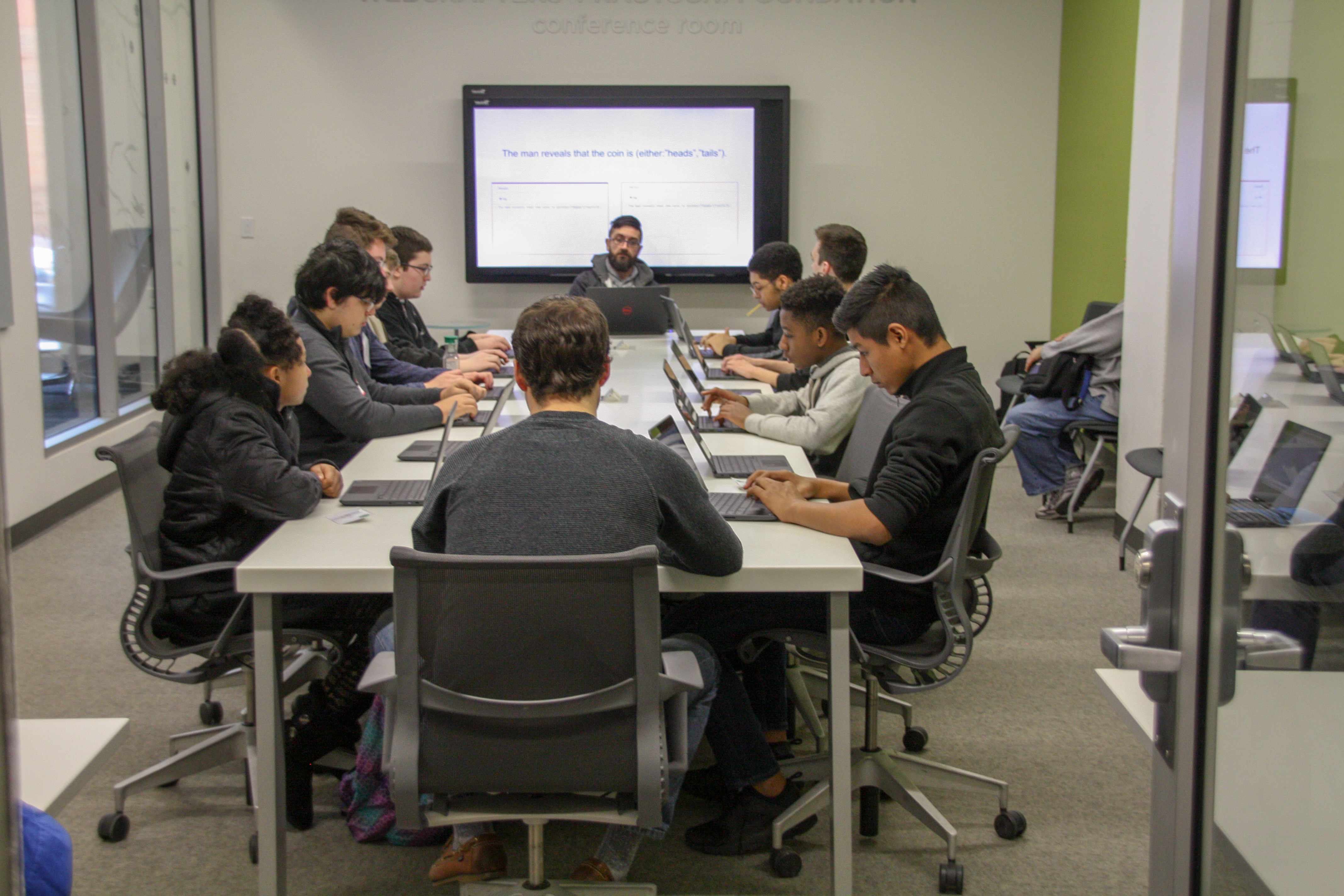Group of your adults with laptopps sitting around a large conference table watching a presentation on a TV
