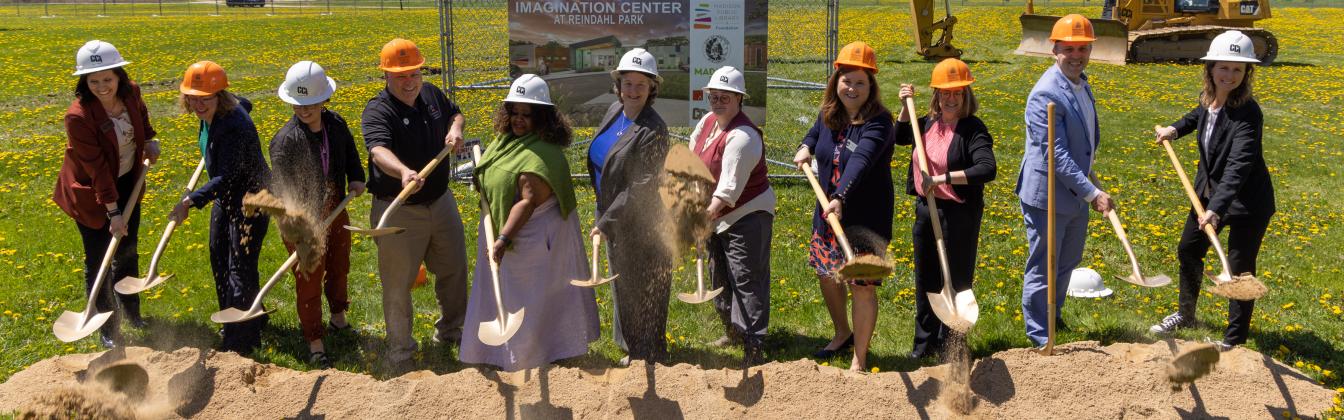 A line of people stand outdoors on a sunny day with hard hats and shovels throwing dirt with construction vehicles behind them