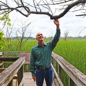 Madison Public Library Naturalist-in-Residence John C. Newman stands among the trees with a prairie in the background at the UW Arboretum