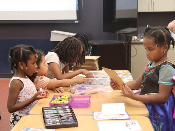 Several young Black girls with braids gather around a table at a library to work on art projects