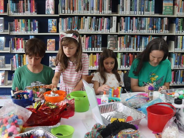 Four kids stand behind a white table full of art supplies working on craft projects in a library