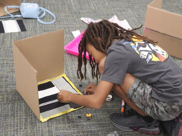 A black child with dreadlocks crouches while working on a diorama made of cardboard