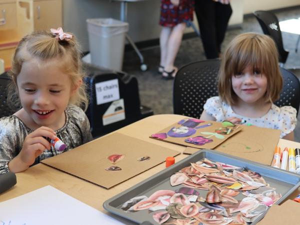 Two young white girls work on paper collages on cardboard while smiling and sitting at a table