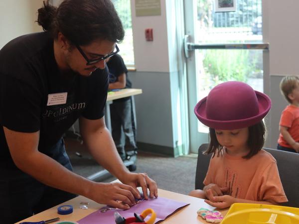 A teaching artist leans over a table to help a little girl with a pink hat with her art project