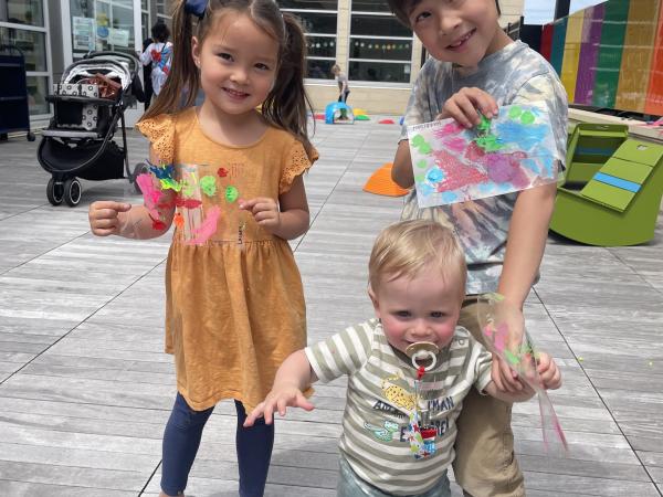 Two older asian kids and a white toddler showcase their art projects outside on a patio at Pinney Library