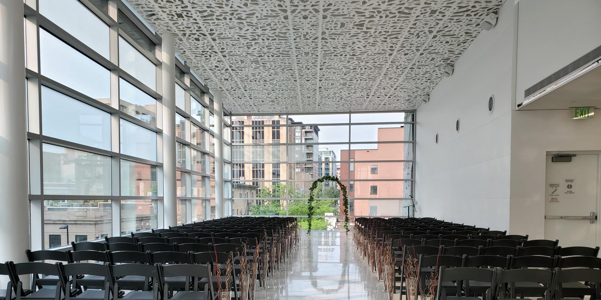 The madison room set up for a wedding ceremony with rows of black chairs and an aisle facing book arch in front of windows