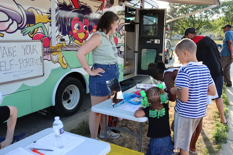 Two kids stand at a table waiting to do their self-portraits in front of a colorful bus and an artist
