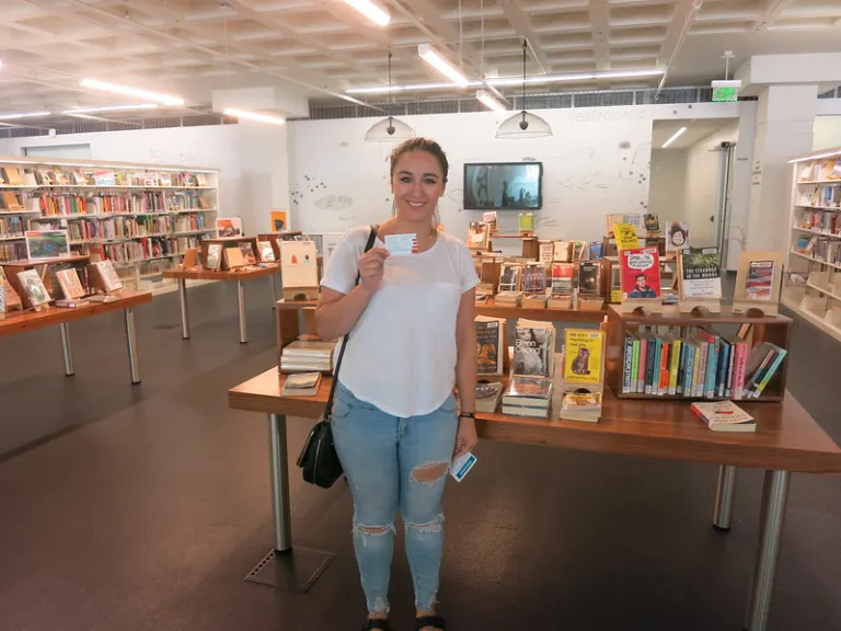 A woman stands holding up a library card in front of a book display at Madison Public Library