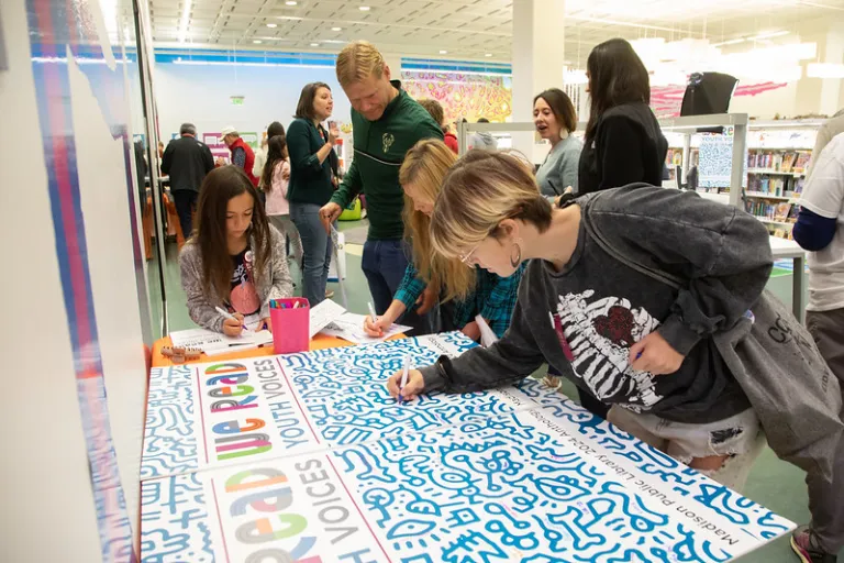 A teen signs a large poster board cover of the WE READ Youth Voices Anthology 2024 as a crowd mingles behind them