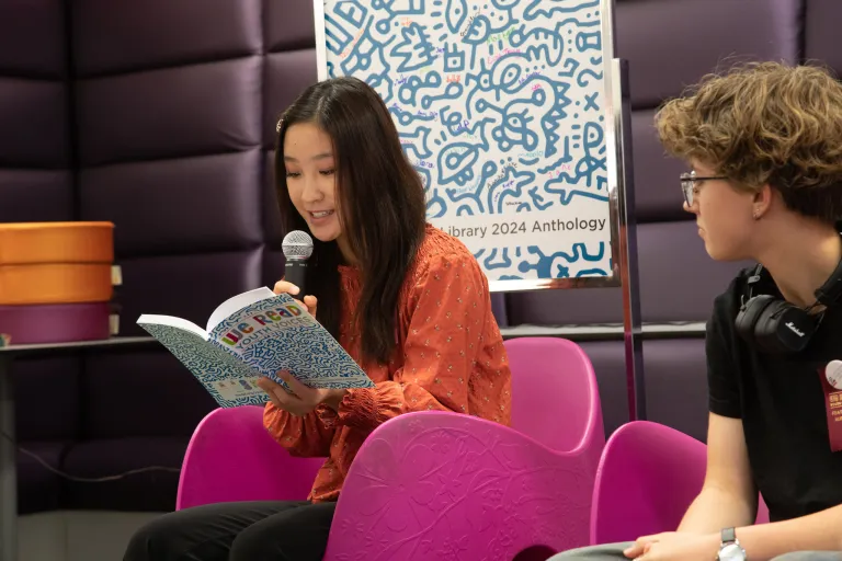 An asian teen reads into a microphone from the WE READ Youth Voices Anthology while seated in a purple chair next to another teen