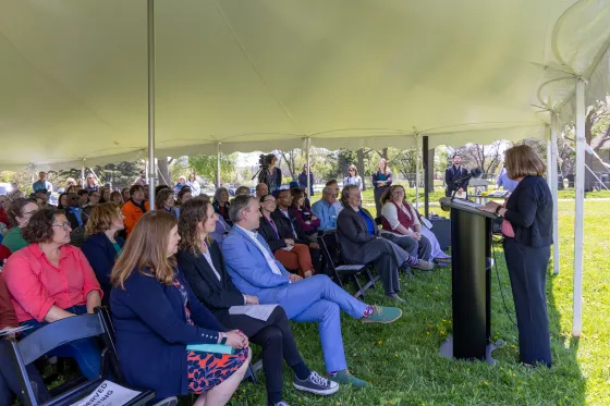 A suited speaker stands at a podium under a tent outdoors and addresses a large, seated crowd