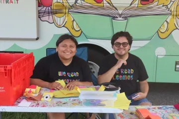 Two Dream Bus employees site behind a low table covered in art supplies in front of Madison Public Library's mobile library