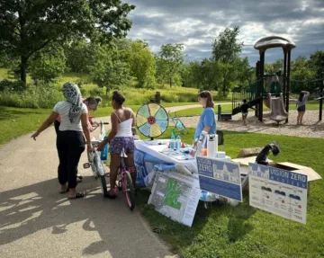 Kids gather around a table staffed by a young woman on a sunny summer day. Yard signs are staked near the table