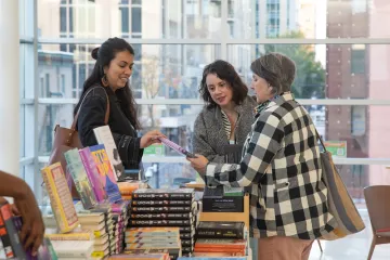Three people stand looking at a book and smiling next to a table full of books and set against a wall of windows